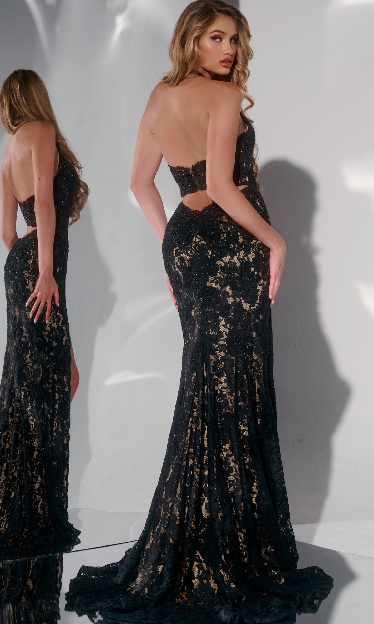 A woman in the Jovani 43097 long prom dress with a black lace corset bodice and nude underlay stands before a mirror, elegantly displaying the gown’s back and side.